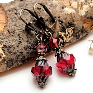 Op de afbeelding: Een paar oorbellen in antieke stijl met een klapbeugel sluiting. De oorbellen hebben gefacetteerde, dieprode glazen kralen en sierlijk metaalwerk in bronskleur. Het ontwerp combineert ronde en geometrische vormen.