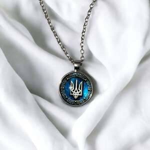 May include: Silver-toned necklace with a round pendant featuring the Ukrainian coat of arms, a trident, set against a blue background. The pendant has a decorative border and hangs from a delicate chain. The necklace is displayed on a white fabric.