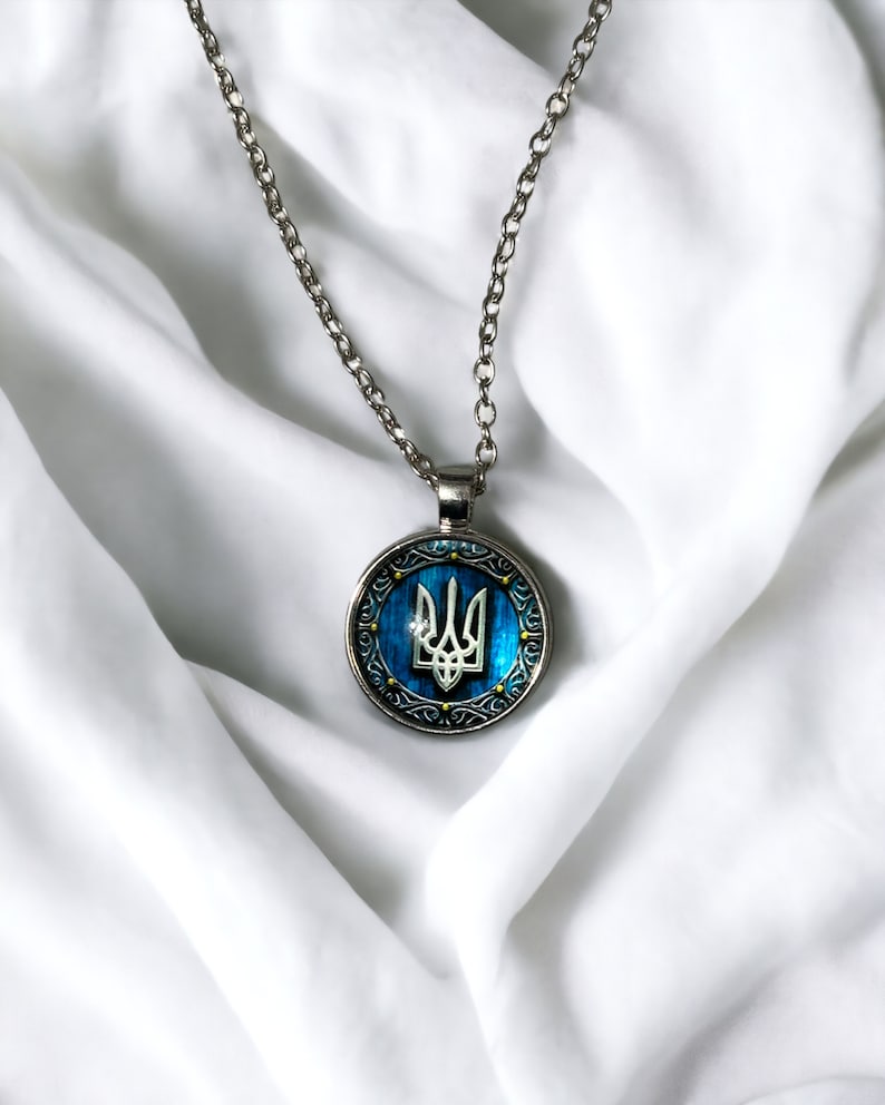 May include: Silver-toned necklace with a round pendant featuring the Ukrainian coat of arms, a trident, set against a blue background. The pendant has a decorative border and hangs from a delicate chain. The necklace is displayed on a white fabric.