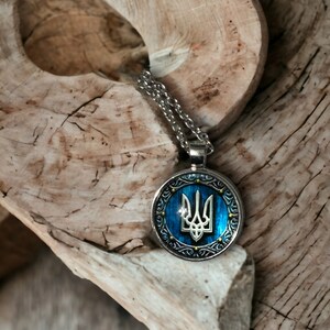 May include: A silver-toned pendant necklace featuring a blue and silver circular pendant with a trident emblem. The pendant hangs on a silver chain and rests on a piece of light-colored wood.