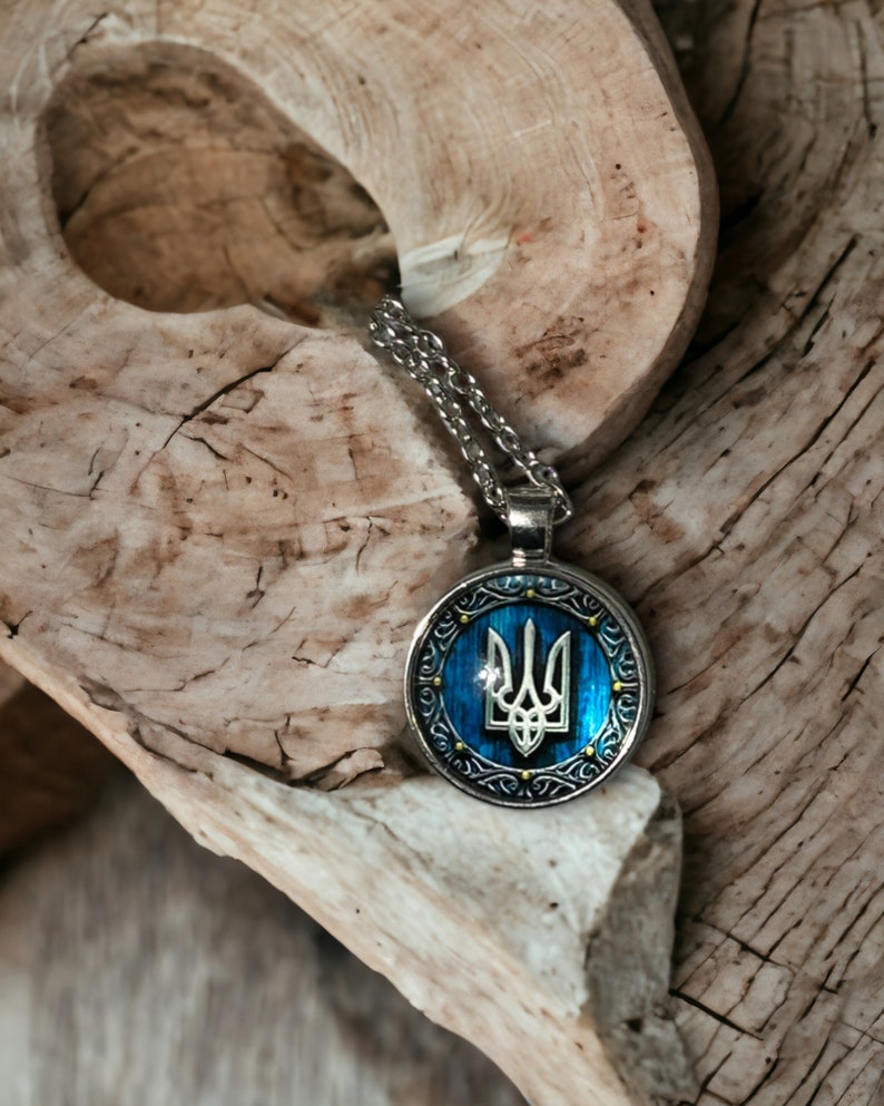 May include: A silver-toned pendant necklace featuring a blue and silver circular pendant with a trident emblem. The pendant hangs on a silver chain and rests on a piece of light-colored wood.