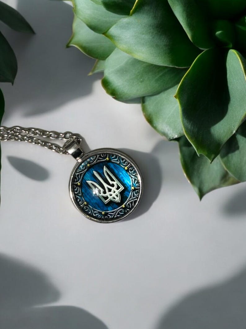 May include: Silver-toned pendant necklace featuring a round pendant with a blue enamel background and a silver trident emblem. The pendant has a decorative border and hangs from a delicate chain. The necklace is displayed on a white surface with green foliage.