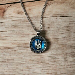 May include: Silver-toned necklace with a round pendant featuring the Ukrainian coat of arms, a trident, on a blue background. The pendant has a decorative silver border with gold accents. The necklace hangs on a wooden surface.