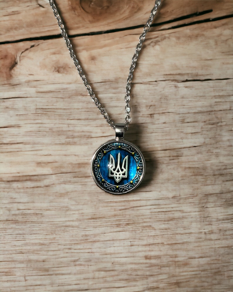 May include: Silver-toned necklace with a round pendant featuring the Ukrainian coat of arms, a trident, on a blue background. The pendant has a decorative silver border with gold accents. The necklace hangs on a wooden surface.