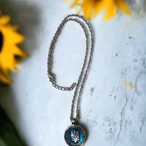 May include: Silver-toned necklace with a round pendant featuring a blue and gold design. The pendant displays a trident symbol, set against a blue background. The necklace is displayed on a white surface with yellow sunflowers in the background.
