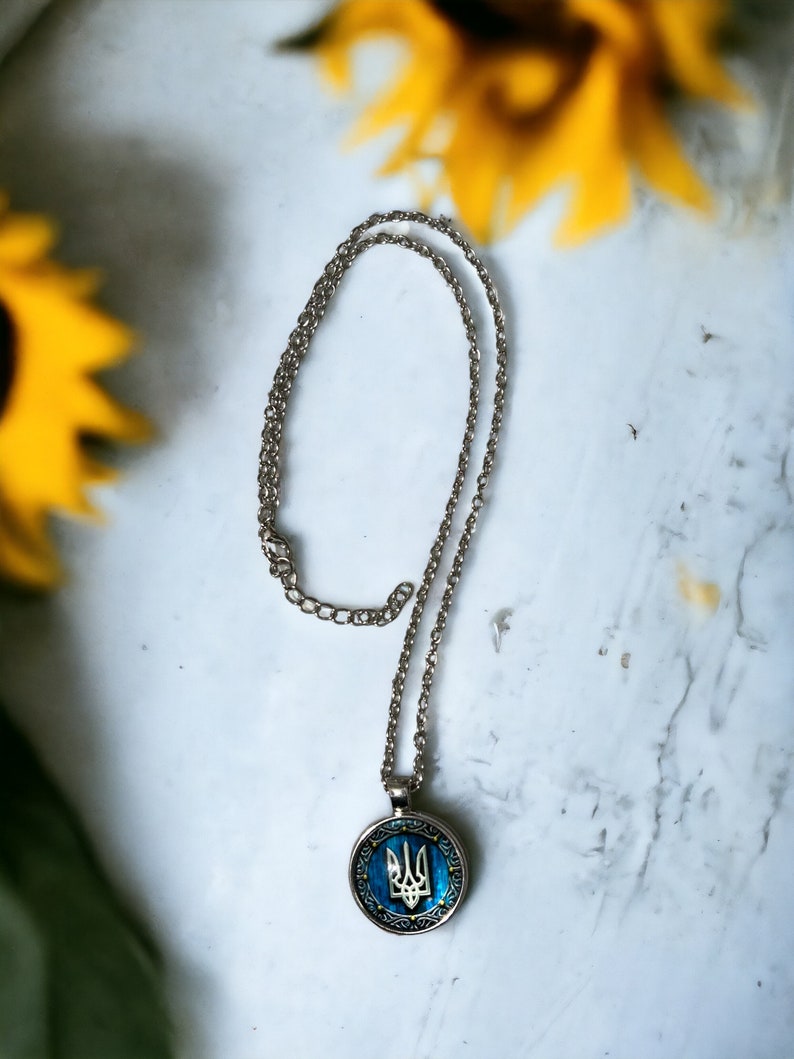 May include: Silver-toned necklace with a round pendant featuring a blue and gold design. The pendant displays a trident symbol, set against a blue background. The necklace is displayed on a white surface with yellow sunflowers in the background.