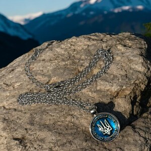 May include: A silver-colored necklace with a round pendant featuring a blue background and a silver trident emblem. The pendant is surrounded by a decorative border. The necklace rests on a textured rock with a mountain backdrop.