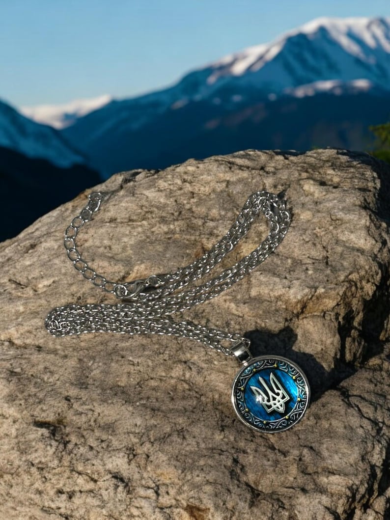 May include: A silver-colored necklace with a round pendant featuring a blue background and a silver trident emblem. The pendant is surrounded by a decorative border. The necklace rests on a textured rock with a mountain backdrop.