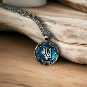 May include: Silver-toned pendant necklace featuring a round pendant with a blue background and a white trident emblem. The pendant is framed by an ornate silver border. The necklace is displayed on a wooden surface.