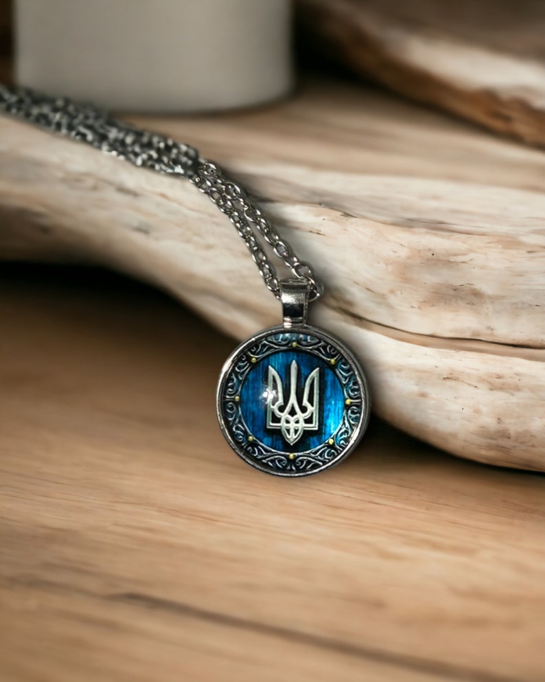 May include: Silver-toned pendant necklace featuring a round pendant with a blue background and a white trident emblem. The pendant is framed by an ornate silver border. The necklace is displayed on a wooden surface.