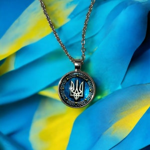 May include: Silver-toned necklace with a round pendant featuring the Ukrainian coat of arms, a trident, in silver against a blue background. The pendant is framed by a decorative silver border. The background is blue and yellow.