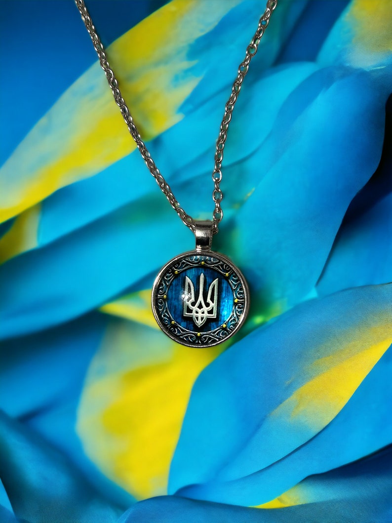 May include: Silver-toned necklace with a round pendant featuring the Ukrainian coat of arms, a trident, in silver against a blue background. The pendant is framed by a decorative silver border. The background is blue and yellow.