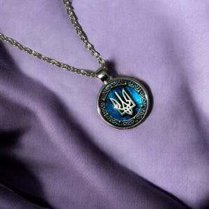May include: Silver-toned necklace with a round pendant featuring the Ukrainian coat of arms, a trident, in silver against a blue background. The pendant is framed by a decorative border. The necklace rests on a purple fabric.