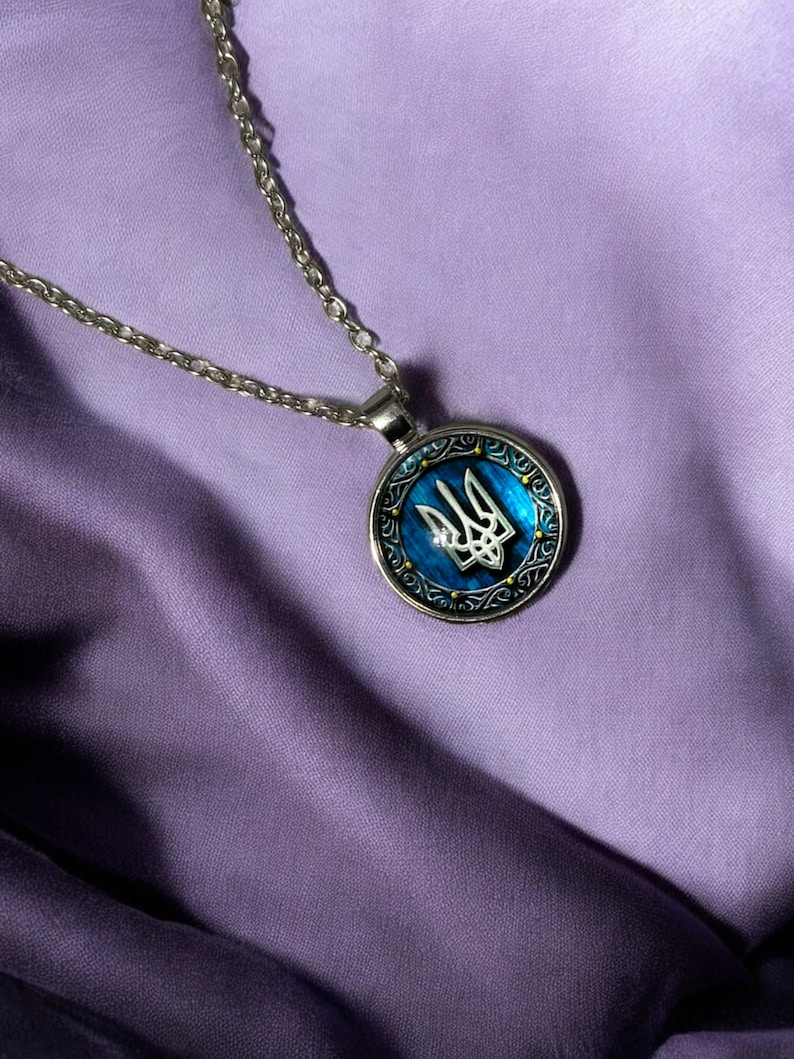 May include: Silver-toned necklace with a round pendant featuring the Ukrainian coat of arms, a trident, in silver against a blue background. The pendant is framed by a decorative border. The necklace rests on a purple fabric.