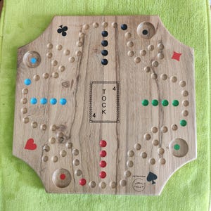 May include: A wooden Tock board game with a natural wood grain finish. The board features four sets of coloured holes and matching game pieces in blue, green, red, and black. The centre is marked with the text TOCK and the number 4 on each side.
