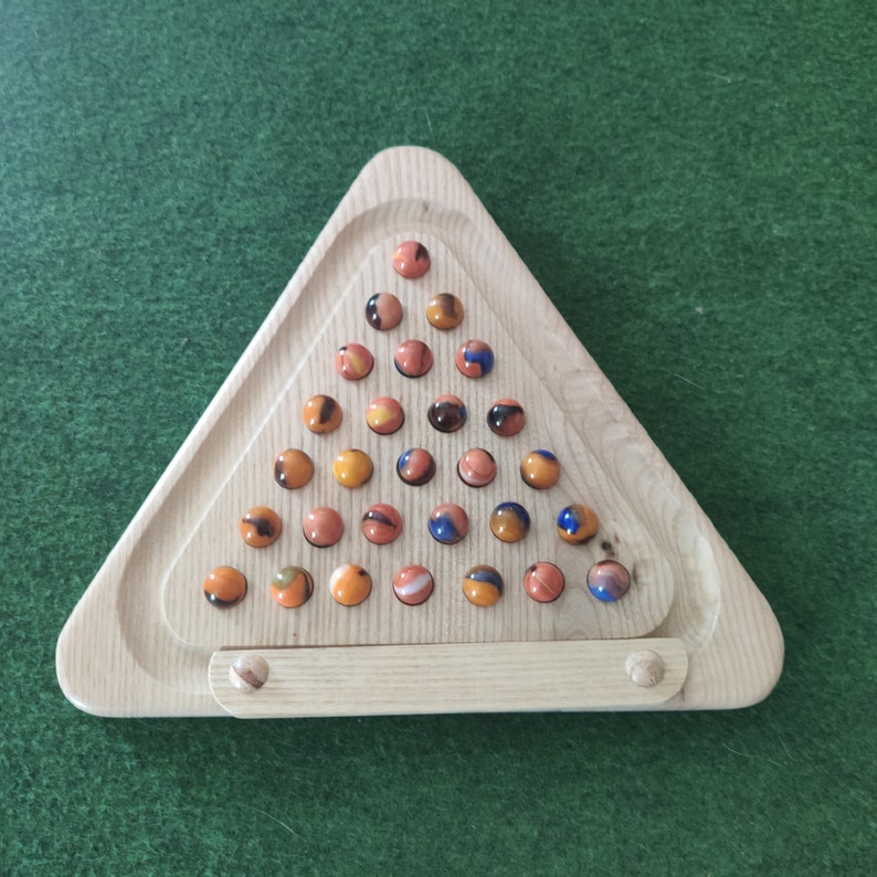 May include: A wooden triangular game board with a marble run. The board is made of light wood and has a triangular shape. The marble run is filled with colorful marbles in shades of orange, blue, and brown.