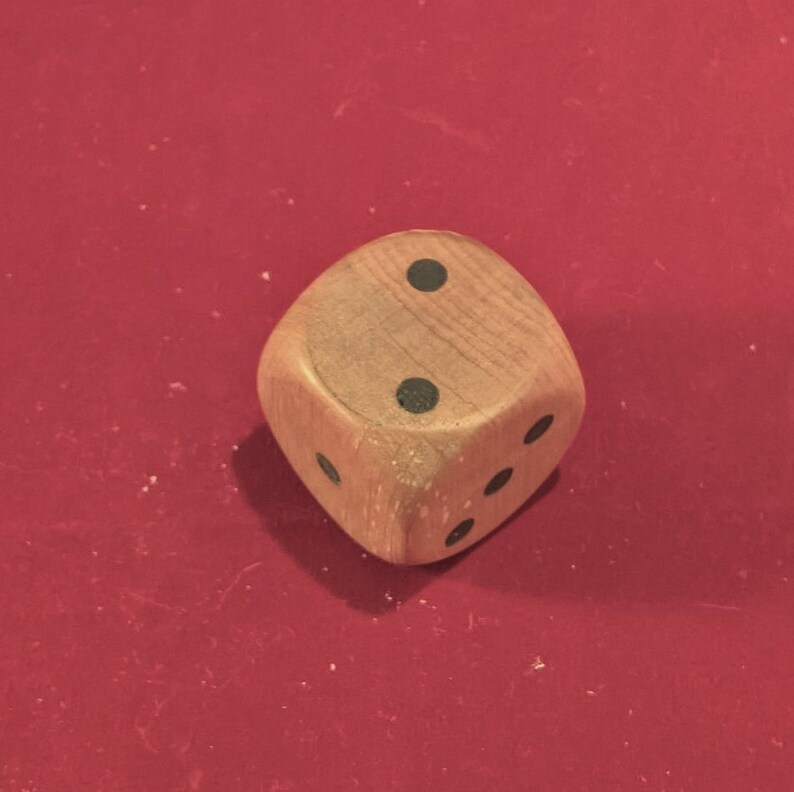 Op de afbeelding: Een houten dobbelsteen met zwarte stippen op elke zijde. De dobbelsteen ligt op een rood oppervlak.