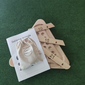 May include: A wooden triangle game with a bag of game pieces. The game board has numbers 10, 15, and 27 printed on it. The game is called "Solitaire Triangle" and is made by "G. Grelet Planete du Jeu".