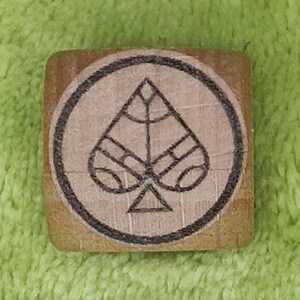 May include: A wooden block with a black circle around a spade playing card symbol. The spade is stylized with a geometric design.