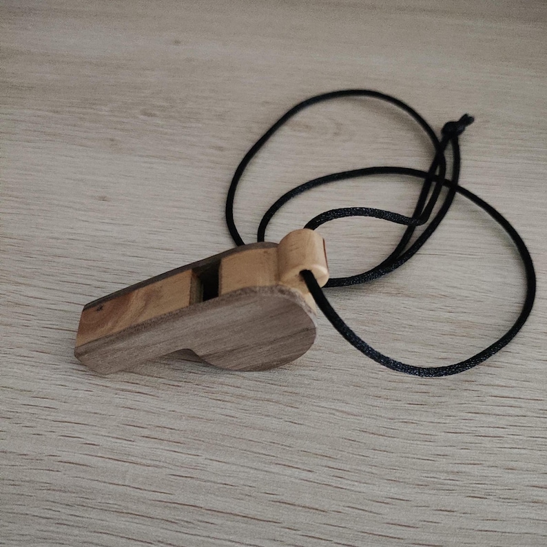 K&ouml;nnte beinhalten: Eine Holzpfeife aus zwei verschiedenen Holzt&ouml;nen mit einem hellen Oberteil und einem dunkleren K&ouml;rper. Die Pfeife ist an einer d&uuml;nnen, schwarzen Kordel befestigt, die eine Schlaufe bildet, sodass sie als Halskette um den Hals getragen werden kann.