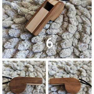 K&ouml;nnte beinhalten: Eine Holzpfeife mit einem geschichteten Design in hellen und dunklen Braunt&ouml;nen. Die Pfeife ist an einer schwarzen Kordel mit einer kleinen hellen Holzperle befestigt. Die Zahl 6 ist in Wei&szlig; auf dem oberen Teil des Bildes aufgedruckt.