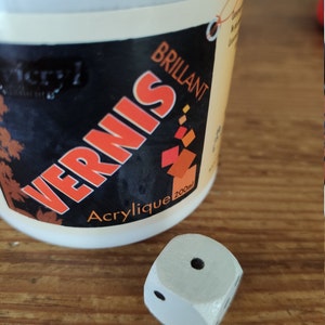 Op de afbeelding: Een witte fles acrylvernis met het label "Vernis Brillant" met een rood en oranje ontwerp. De fles is 200 ml en staat op een houten oppervlak naast een witte houten dobbelsteen met zwarte stippen.