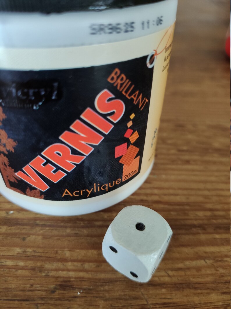 Op de afbeelding: Een witte fles acrylvernis met het label "Vernis Brillant" met een rood en oranje ontwerp. De fles is 200 ml en staat op een houten oppervlak naast een witte houten dobbelsteen met zwarte stippen.