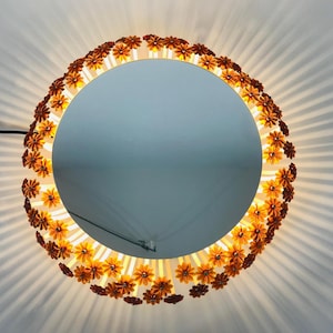 Op de afbeelding: Een ronde spiegel met een decoratieve bloemenrand. De rand is versierd met kleine, oranje en bruine bloemen die in een cirkel zijn gerangschikt. De spiegel wordt verlicht met een warm licht.