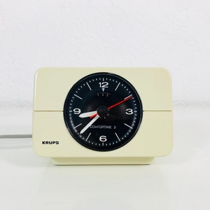 May include: A white analog alarm clock with a black face and black hands. The clock has the text "COMFORTIME 3" and "KRUPS" printed on it.