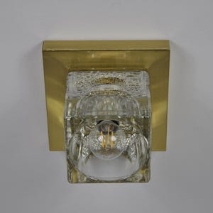 May include: A vintage wall sconce featuring a clear, textured glass shade and a gold-coloured metal base. The light fitting has a single bulb visible inside the glass. The sconce is mounted on a white wall.