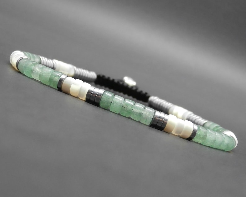 May include: A green and white beaded bracelet with a black cord. The bracelet features green jade beads, white shell beads, and silver metal beads.