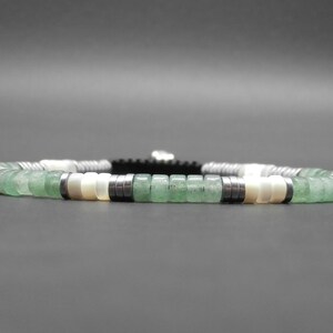 May include: A green and white beaded bracelet with silver accents. The bracelet is made with round beads and has a black cord.