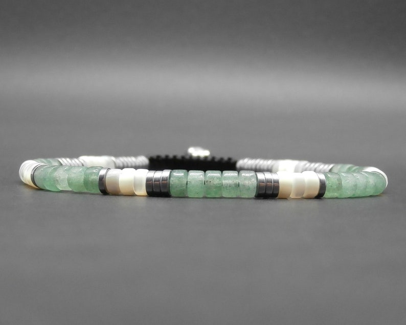 May include: A green and white beaded bracelet with silver accents. The bracelet is made with round beads and has a black cord.