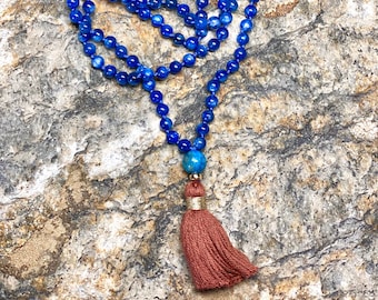 May include: A blue gemstone mala necklace with a tassel. The necklace is made of round beads and has a brown tassel.