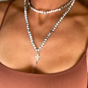May include: A layered necklace with a cross pendant. The top layer is a white beaded necklace, and the bottom layer is a blue and brown beaded necklace with a silver cross pendant.
