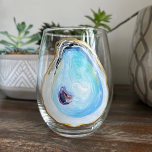 Könnte beinhalten: Ein klares, stieloses Weinglas mit einem handgemalten Austerndesign. Die Auster ist in Blau-, Weiß- und Lilatönen mit einer goldenen Umrandung bemalt. Das Glas steht auf einer Holzoberfläche.