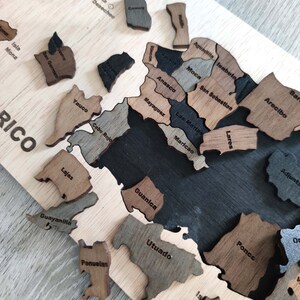 May include: A wooden puzzle map of Puerto Rico with the names of each municipality labeled. The puzzle pieces are in various shades of brown and black.