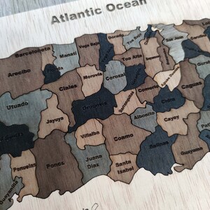 May include: A wooden map puzzle of Puerto Rico with the names of the municipalities labeled on each piece. The puzzle is shaped like the island and is surrounded by a light brown border. The text "Atlantic Ocean" is at the top of the puzzle. The text "Isla Caja de Muertos" is at the bottom of the puzzle.