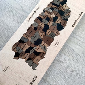 May include: A wooden map of Puerto Rico with the names of each municipality labeled. The map is divided into different shades of brown and black. The map is surrounded by a light brown wooden frame. The words "Puerto Rico" are written in large, bold letters at the bottom of the map. The words "Atlantic Ocean" are written at the top left of the map. The words "Caribbean Sea" are written at the top right of the map.