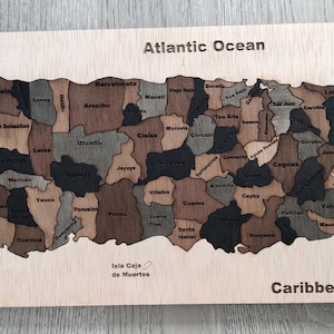 May include: A wooden puzzle map of Puerto Rico, showing the different municipalities of the island. The map is labeled with the names of the municipalities and the surrounding bodies of water, including the Atlantic Ocean and the Caribbean Sea.