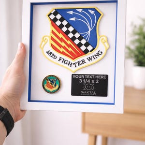 May include: Framed military emblem featuring a shield with red, yellow, blue, and black accents. The frame includes a small coin and a black plaque with the text "YOUR TEXT HERE 3 1/4 x 2". The emblem is labeled "482D FIGHTER WING".
