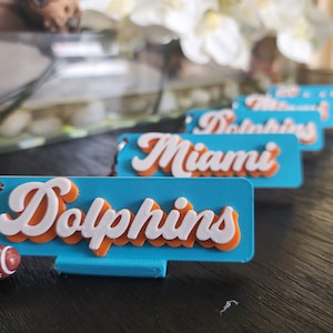 Puede incluir: Cinco llaveros azul turquesa con letras blancas y naranjas. Los llaveros deletrean "Dolphins" y "Miami". Un llavero tiene un pequeño amuleto de fútbol marrón y blanco.