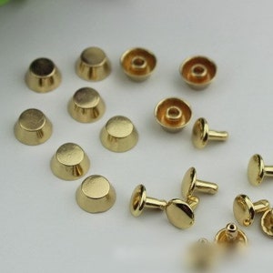 May include: A collection of gold-toned metal rivets and studs, some with a domed top and others with a flat top. The rivets have a post on the bottom for attaching to fabric or leather.