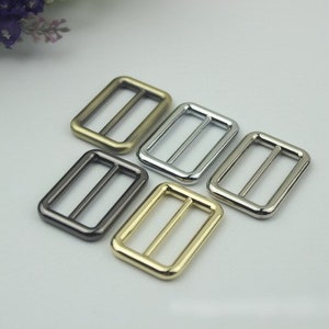 Könnte beinhalten: Vier Metall-Schiebeschließen in verschiedenen Ausführungen: Gold, Silber, Schwarz und Messing. Die Schließen sind rechteckig mit einem Mittelbalken.