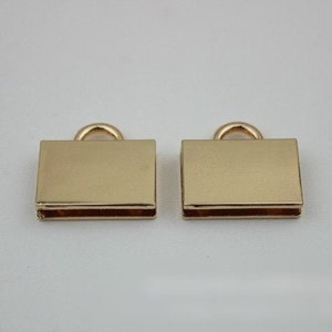 May include: Two gold-colored metal clasps with a loop on top. The clasps are rectangular and have a smooth, shiny finish.