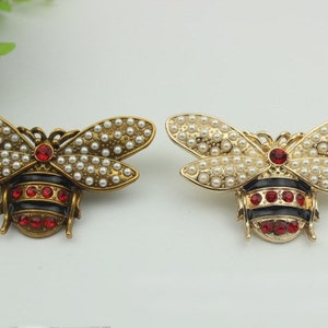 May include: Two gold bee brooches with black and red enamel details. Each bee is decorated with white pearl-like beads.