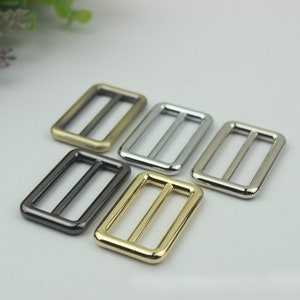 Könnte beinhalten: Fünf Metall-Schiebeschließen in verschiedenen Ausführungen: Gold, Silber, Schwarznickel und zwei Brauntönen. Die Schließen sind rechteckig mit einer einzigen, mittigen Öffnung.