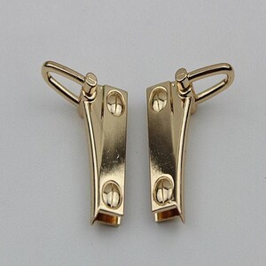 May include: Two gold-toned metal clasps with a loop on each side. The clasps have a curved shape and four small, round, raised details on each side.