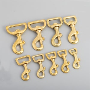 May include: A collection of brass-colored swivel snap hooks in various sizes. The hooks have a rectangular or D-shaped loop at the top and a spring-loaded clasp at the bottom. The hardware is arranged on a light gray surface.