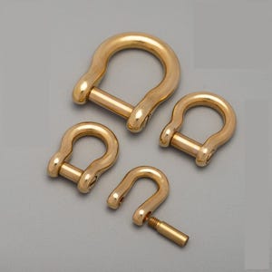 Könnte beinhalten: Vier goldfarbene D-Schäkel in verschiedenen Größen, angeordnet auf einer grauen Oberfläche. Die Schäkel haben eine U-Form mit Stiftverschluss. Ein Schäkel hat seinen Stift teilweise abgeschraubt. Das Metall hat eine polierte Oberfläche.